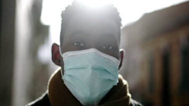 Siyahi bir adam güneş ışığında dışarıda kameraya bakarak maske takıyor.