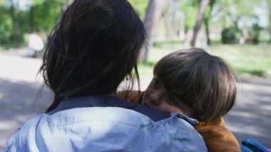 Anne oğlunu yanağından öpüyor, park bankında oturuyor. Yorgun çocuk öğle uykusundan uyandı. Ebeveyn çocuğu kucaklıyor.