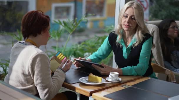 コーヒーショップに座っている女性スタッフにタブレットを見せるビジネスウーマンマネージャー 女性起業家は新しい従業員に仕事を説明する ノートを書く女は指示を学ぶ — ストック動画