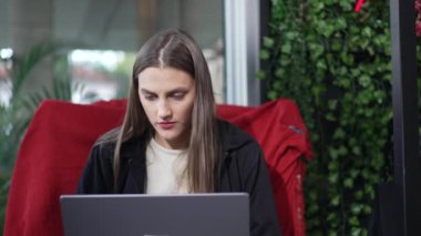 Laptop teknolojisi kullanan genç bir kadın kahve dükkanından uzaktan çalışıyor. Bilgisayar ekranının önünde oturan 20 'li yaşlardaki kız kırmızı koltukta oturuyor.