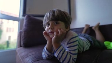 Mutlu küçük çocuk evinde kanepede uzanmış, çenesi çenesinde kamera karşısında gülümsüyor.