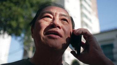 Happy senior Asian man speaking on phone while walking in city street. Portrait of an older middle aged person talking in conversation with cellphone device