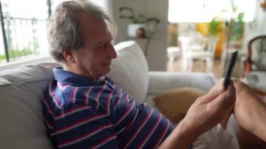 70 'li yaşlarda elinde akıllı telefon cihazı olan olgun bir adam. Üst düzey kişi oturma odasındaki koltukta oturan aile üyesiyle konuşmak için modern teknolojiyi kullanıyor.
