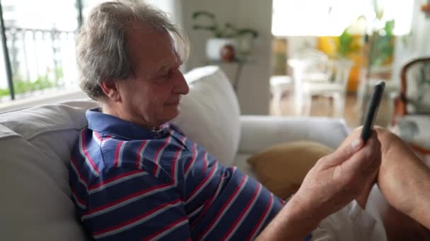 70代の熟年男性がスマートフォンを持っています リビングルームのソファに座って家族と話すために現代的な技術を使用してシニア人 — ストック動画