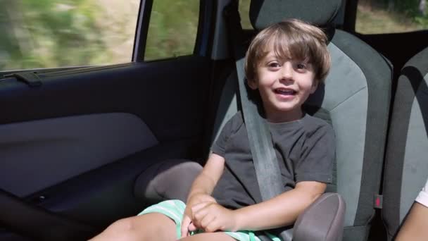 一个快乐的小男孩坐在车后座上对着摄像机笑 带安全带在路上行走的儿童 — 图库视频影像