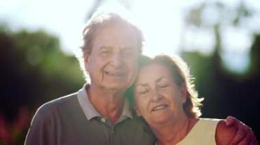 Yaşlı çift parkta kameraya gülümsüyor. Yaşlı karısına sarılmış bir kocanın portresi. Arkaplan ışığı