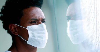 Black African man wearing virus mask looking out window, glass face reflection