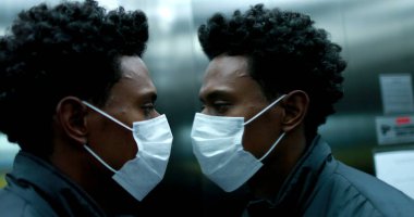 Black african man wearing face mask looking at himself in elevator mirror