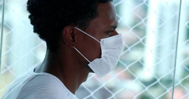 Siyah koronavius yüz maskesi takan Afrikalı bir adam evde pencereden dışarı bakıyor.