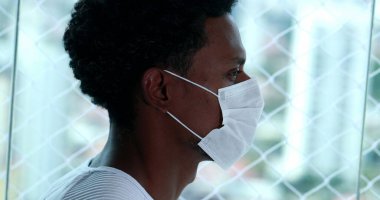 Siyah koronavius yüz maskesi takan Afrikalı bir adam evde pencereden dışarı bakıyor.