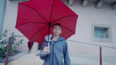 Yağmur yağarken elinde kırmızı şemsiyeyle dışarıda yürüyen genç bir çocuk. Ergen çocuk yağmurlu bir günde dışarı çıkar.