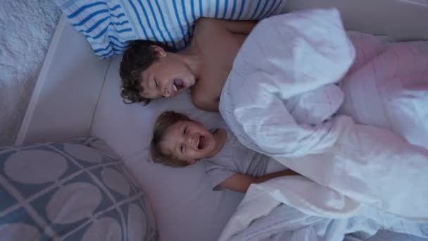 快乐的两个兄弟在床单下 两个弟弟妹妹躺在床上笑着 真正的家庭时刻 孩子们从床单上偷看 — 图库视频影像