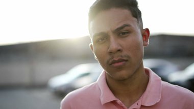 Ciddi bir ifadeyle kameraya bakan şüpheci İspanyol bir gencin portresi. Güney Amerikalı Brezilyalı erkek güneş ışığında dışarıda duruyor.