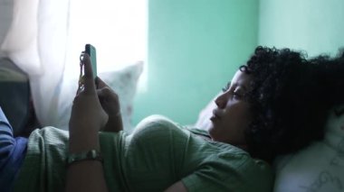 Yatakta uzanmış telefonu kullanan siyah bir kadın. Akıllı telefona bakan Afrikalı bir Amerikalı.