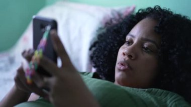 Yatakta uzanmış cep telefonu cihazına bakan genç bir zenci kadın.