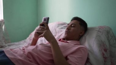 Gizli bir Güney Amerikalı genç adam akıllı telefon cihazıyla yatakta yatıyor.