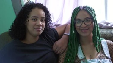 İki siyah İspanyol kadın koltukta oturmuş kameraya bakıp gülümsüyor.
