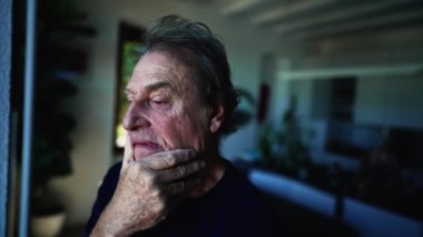 Düşünceli bir ifadeyle pencerenin kenarında duran son sınıf öğrencisi. 70 'li yaşlarda evde oturan olgun bir adamın endişeli ifadesi. kaygılı duygu.