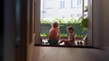 Çocuklar kapının önünde oturuyor. Ayakkabılarını dışarıda giyiyorlar. Büyük kardeş, küçük kardeşe ayakkabıyı kapının önüne koyması için yardım ediyor.