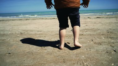 Çocuk kumsalda çıplak ayakla yürüyor pantolonu sarılmış, kumların çıplak ayakla yürüdüğünü hissediyor.