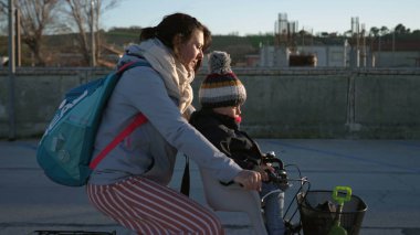 Anne, soğuk kış gününde arka planda güneş ışığı parlarken çocukla birlikte bisiklete biner. Annem bisiklete biniyor