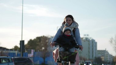 Anne deniz kıyısındaki bisiklet yolunda çocuğuyla bisiklet sürüyor. Soğuk bir günde ebeveyn bisiklete biniyor.