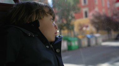 Düşünceli çocuk otobüs penceresinin önünde oturmuş hareketli şehre bakıyor. Düşünceli küçük bir çocuk toplu taşımada cama yaslanıyor.