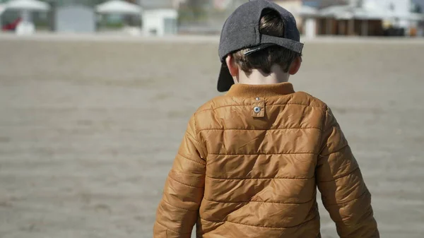 Back of child walking at beach wearing winter jacket and cap