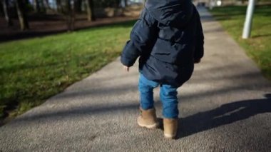 Güneşli kış gününde parkta yürürken ceketi ve botları olan küçük bir çocuk.