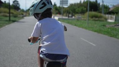 Yeşil yolda bisiklet süren bir çocuğun arkası. Bir çocuk güneşli bir günde dışarıda kask takarak bisiklet sürüyor. Çocukluk egzersizi.