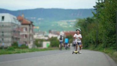 Yeşil yolda üç tekerlekli scooter süren neşeli küçük bir çocuk. Çocuk, güneşli bir günde kaskıyla dışarıda oyuncak scooter sürüyor.
