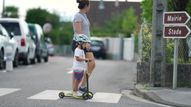 Anne ve çocuk kaldırımda karşıdan karşıya geçiyorlar. Miğfer takan çocuk zebra çizgilerinde üç tekerlekli scooter kullanıyor.