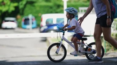 Kaldırımda bisiklet süren bir çocuk. Koruyucu kask ve tekerlek desteği takan genç bir çocuk şehir caddesinde annesiyle bisiklete biniyor.