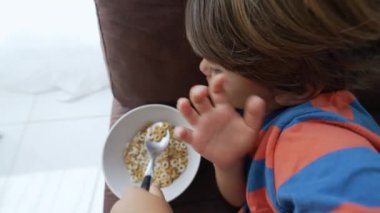 Çocuk kanepede uzanmış mısır gevreği yiyor. Küçük bir çocuk kaşıkla sabah yemeği yiyor.