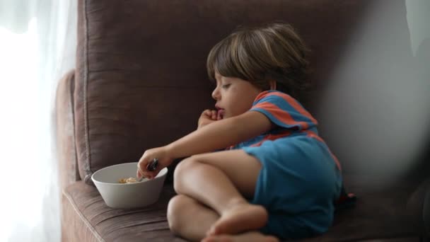 子供がソファに寝そべってシリアルを食べるなんて 子供はスプーンでソファで朝の朝食を食べます 週末を楽しむリラックスした子供 — ストック動画