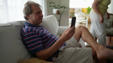 Telefonu elinde tutan kıdemli adam akıllı telefonla konuşuyor. Sıradan, otantik, olgun bir insan oturma odasındaki koltukta modern teknoloji cihazı kullanıyor.