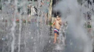  Küçük çocuk yaz günü şehir parkında su jetlerinde koşuyor. Küçük çocuk ferahlatıcı suda koşuyor yavaş çekimde sıçrıyor