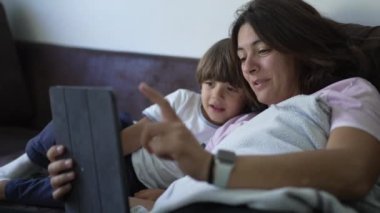 Mutlu gizli anne ve küçük oğlu evde kanepede tablet kullanıyor. Çocuk ve anne birlikte ekrana bakıp gülümsüyor.