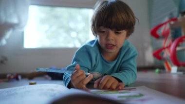 Yoğunlaşmış bir çocuk yatak odasında ödevlerini pijama giyerek yapıyor. Çocuk elinde renkli kalemle yerde yatıyor.