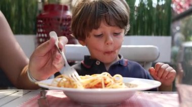 Yemek masasında makarna yiyen bir çocuk. Ebeveyn tabakta domates soslu spagetti alıyor. Restoran yemeğindeki genç çocuğun yüzü.