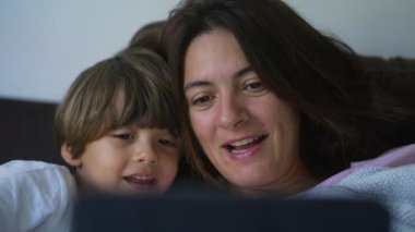 Anne ve çocuk yüzlerini kapatıp birlikte kanepede uzanmış tablet ekrana bakıyorlar.