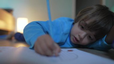 Çocuk renkli kalemle kağıda resim çiziyor. Çocukluk yaratıcı aktiviteleri. Anaokulu öğrencisi evde yerde yatıyor.