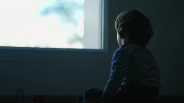 Karanlıkta oturan yalnız küçük bir çocuk pencereye bakıyor. Üzgün çocuk yatak odasında yalnız.