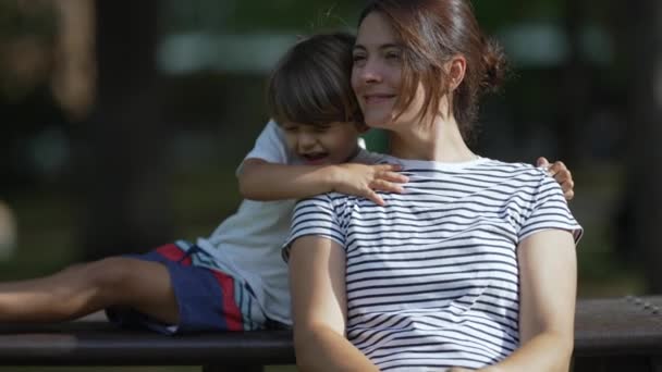 儿子抱着妈妈一起坐在公园的长椅上 在美丽的阳光明媚的日子里 妈妈和孩子们拥抱大自然 — 图库视频影像