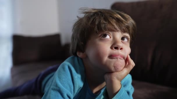 小さな男の子が集中的な表現と顎の手でソファの上に横たわっています 家のソファでパジャマを着た思慮深い子供の肖像画 — ストック動画