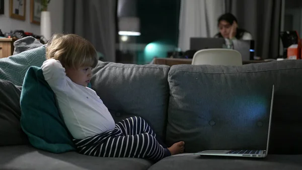 Child watching computer laptop screen while mother works at night