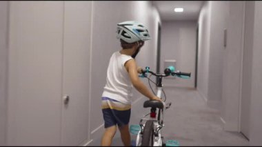 Bir çocuk bina koridorunda bisiklet taşıyor ve asansör düğmesine basıyor. Koruyucu kask takan çocuk hafta sonu etkinliğine hazırlanıyor..