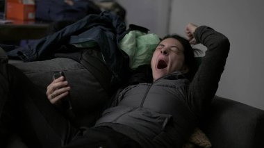 Tired woman lying down in sofa yawning and talking on phone with family