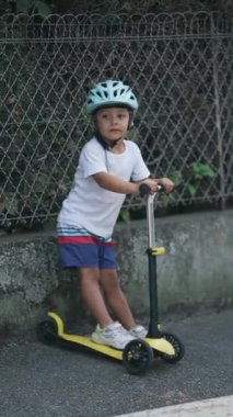 Üç tekerlekli scooterlı ve koruyucu kasklı, sokakta kameraya poz veren sevimli küçük çocuk.