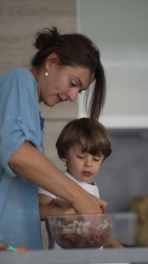 Anne, malzemeleri çocukla karıştırıp hamuru yiyor. Ebeveyn ve genç çocuk dikey videoda birlikte yemek hazırlıyor ve pişiriyorlar.